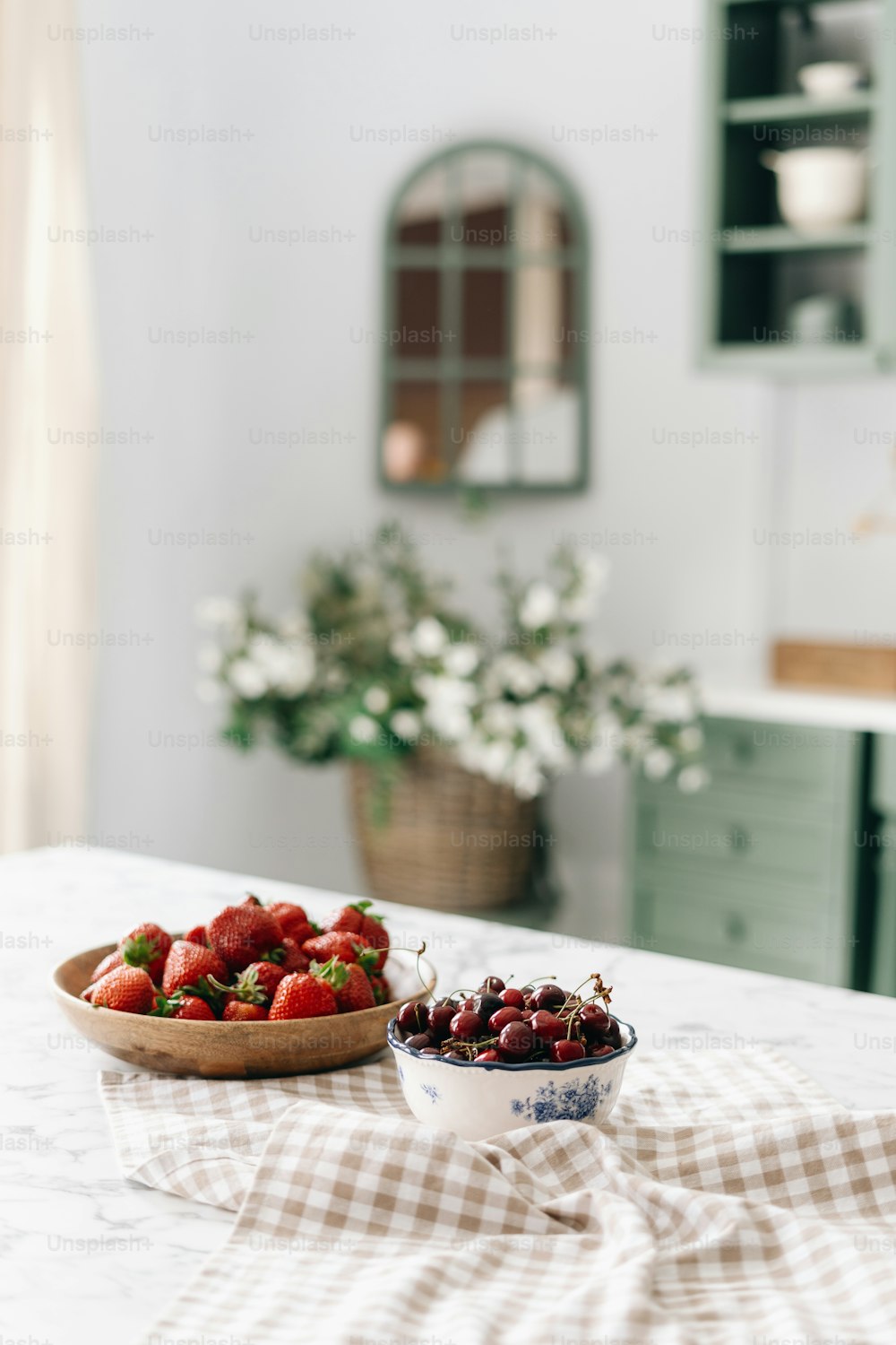 Pair of bowls full of cherries and strawberries on table cloth on kitchen island with marble top,, green vintage furniture in blurred background, woven basket filled with flowers. Vertical shot