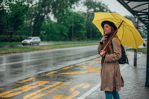 Junge schöne Frau, die einen gelben Regenschirm hält und an einem regnerischen Tag an einer Bushaltestelle auf ein öffentliches Verkehrsmittel wartet