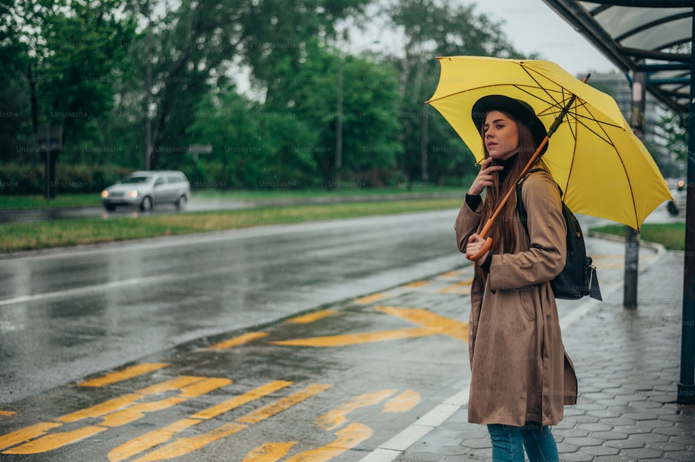 Young beautiful woman holding a yellow umbrella and standing on a bus stop while waiting for a public transport on a rainy day