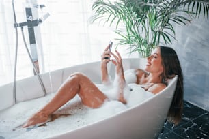 Uses phone. Beautiful young woman have free time at home in the bath.