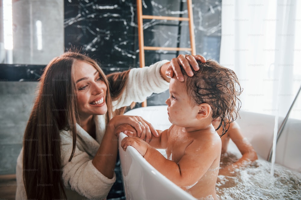若い母親は息子と娘を助けます。お風呂で体を洗う2人の子供。