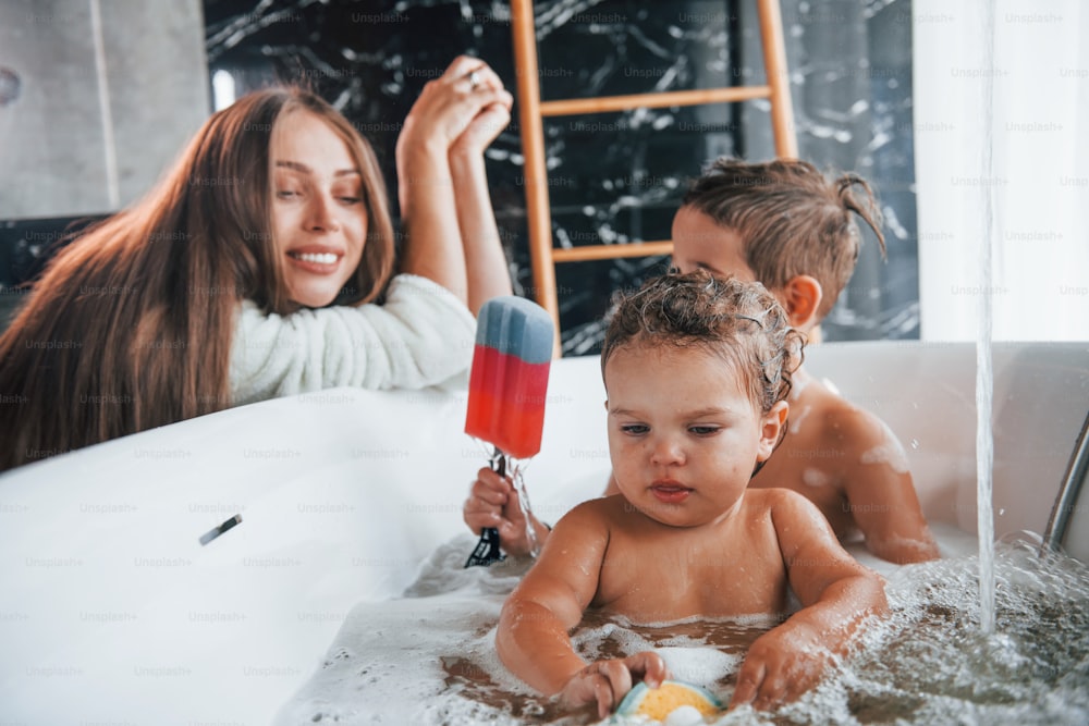 A jovem mãe ajuda o filho e a filha. Duas crianças lavando no banho.