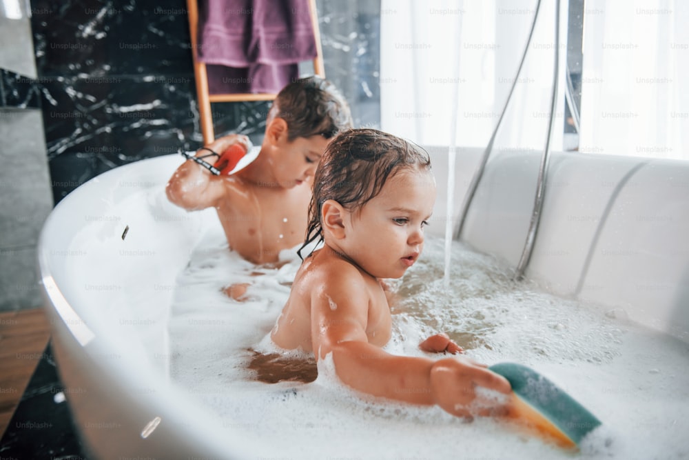 Two kids having fun and washing themselves in the bath at home. Helping each other.