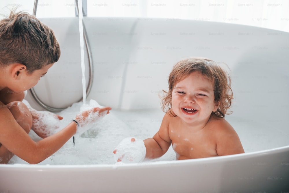 Two kids having fun and washing themselves in the bath at home. Posing for a camera.