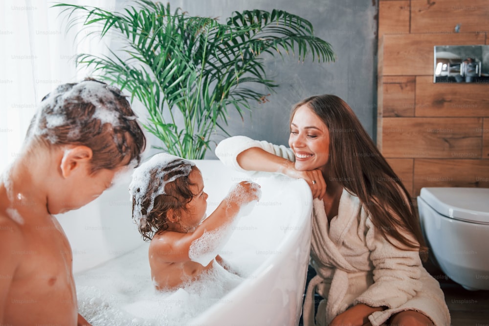 Divertindo-se. A jovem mãe ajuda o filho e a filha. Duas crianças lavando no banho.
