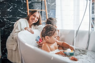 Young mother helps her son and daughter. Two kids washing in the bath.