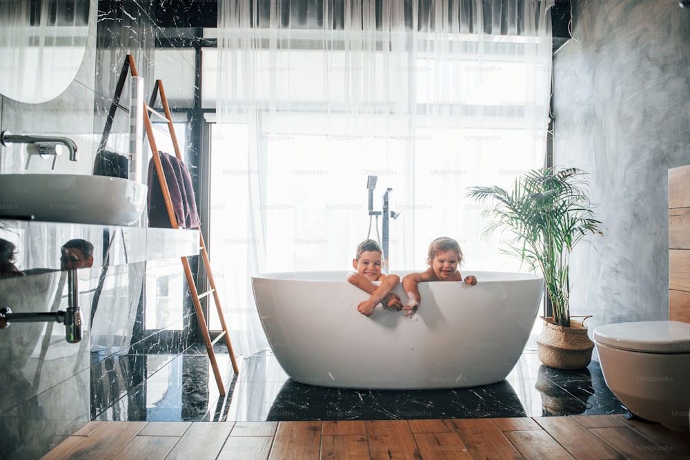 Two kids having fun and washing themselves in the bath at home. Posing for a camera.