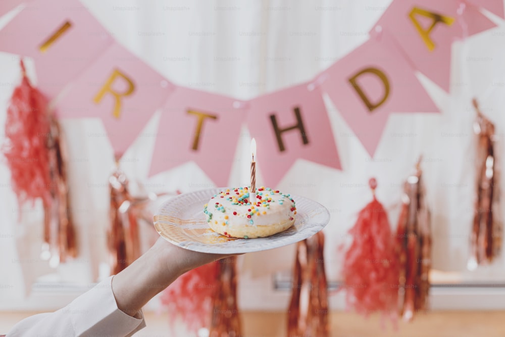 Delicious birthday donut with candle in hand on background of pink garland and decorations in festive room. Celebrating birthday party. Colorful doughnut with sprinkles and rose gold candle