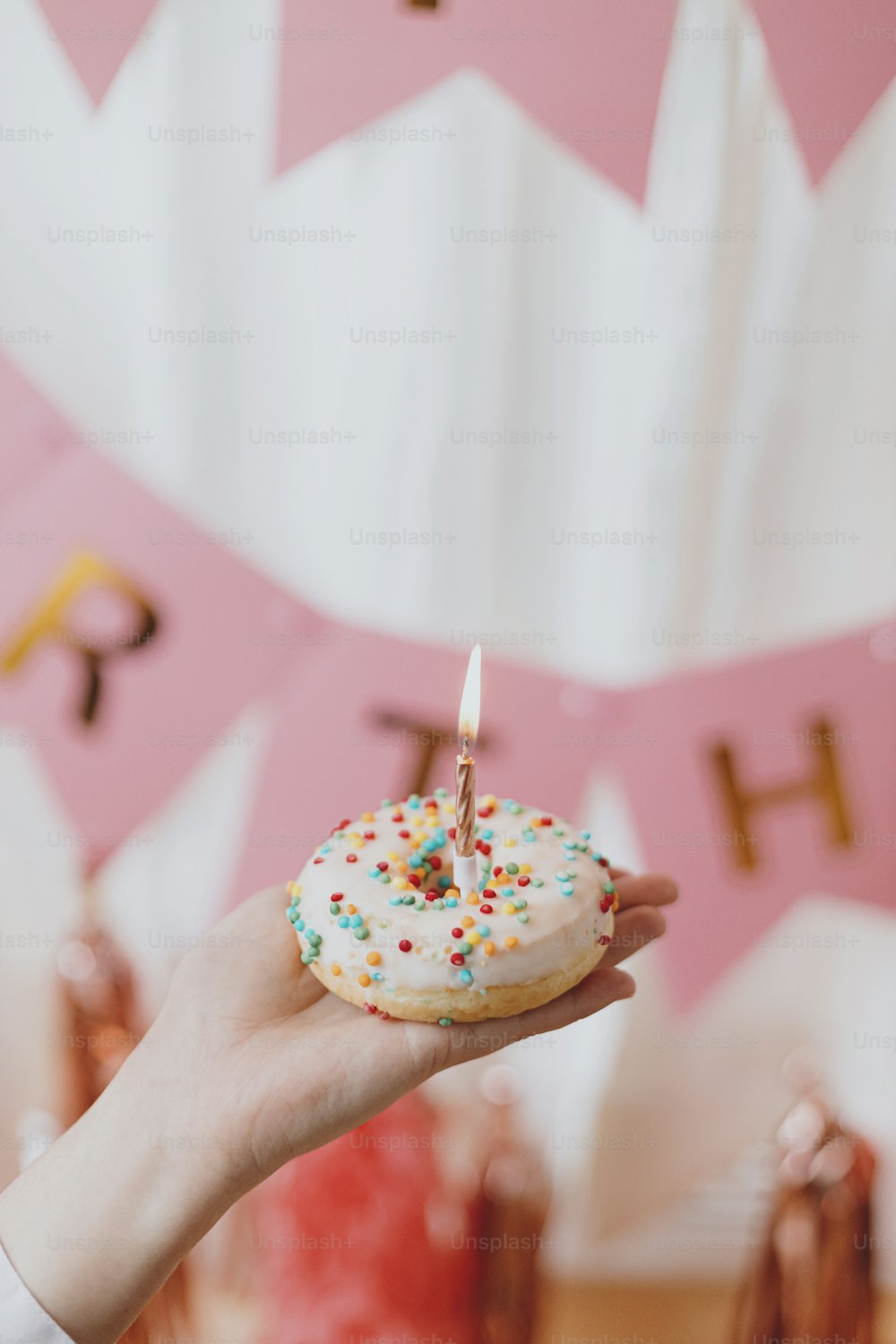 Delicious birthday donut with candle in hand on background of pink garland and decorations in festive room. Celebrating birthday party. Colorful doughnut with sprinkles and rose gold candle