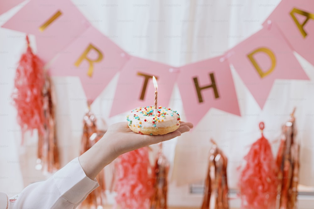 Happy Birthday. Hand holding delicious birthday donut with candle on background of pink garland and decorations in room.  Birthday party celebration. Colorful doughnut with sprinkles