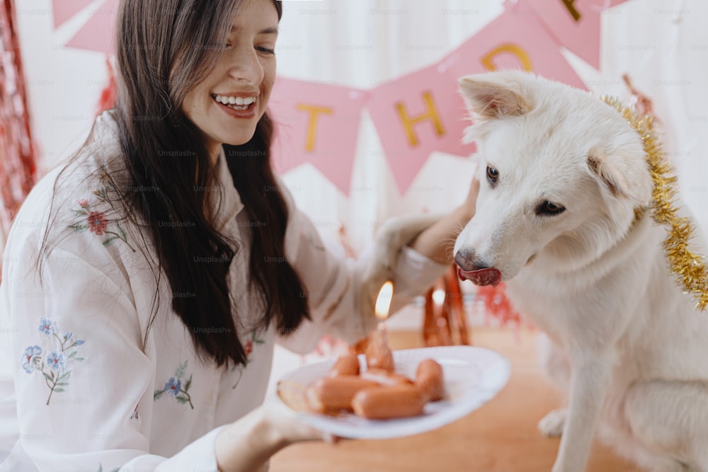 Dog birthday party. Happy young woman and hungry dog celebrating birthday with sausage cake and candle on background of pink garland. Adorable white swiss shepherd dog first birthday