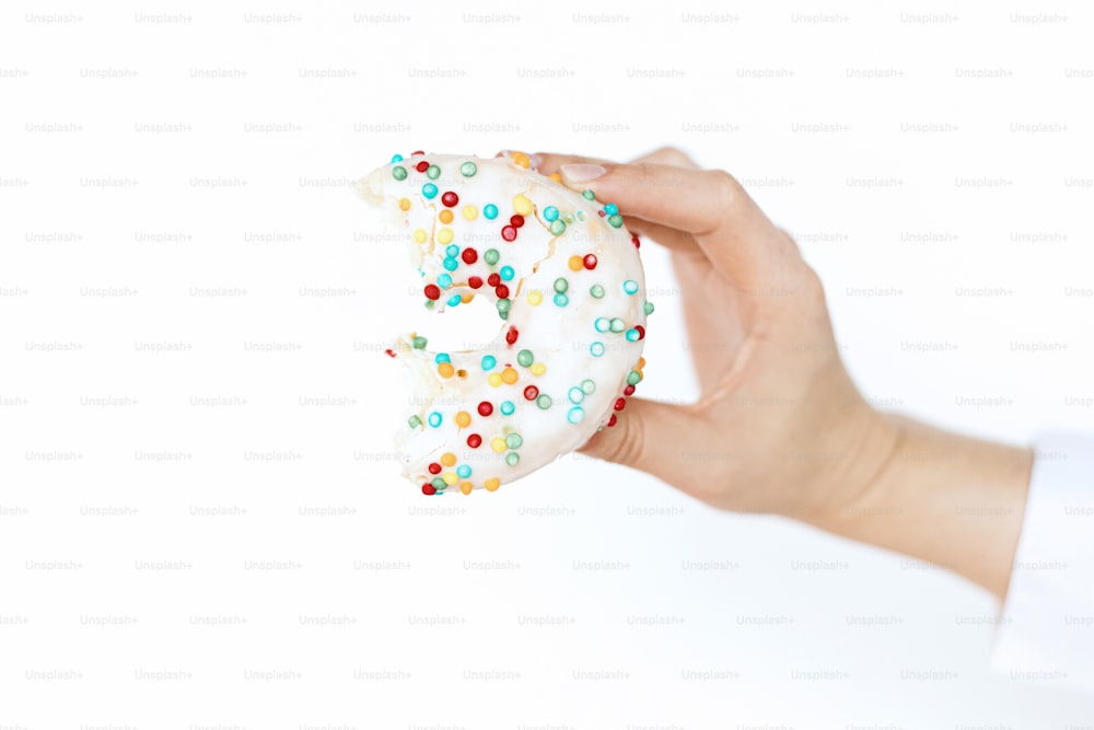 Delicious bitten donut in hand on background of white wall in room. Celebrating birthday party. Colorful doughnut with sprinkles. Diet and moment of happiness