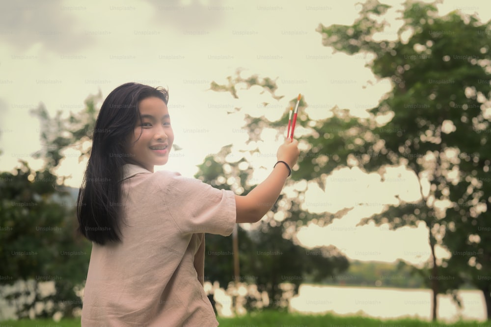 Asian girl holding painting brush and smiling to camera while standing in the park.