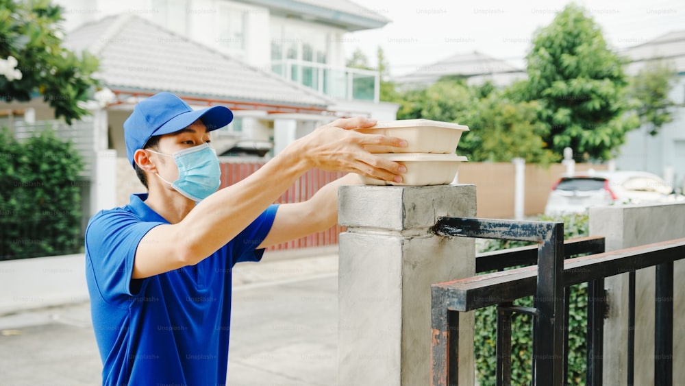 Le jeune homme de courrier de livraison postale porte un masque facial manipulant la boîte de nourriture pour envoyer au client à la maison et la femme asiatique reçoit le colis livré à l’extérieur. Mode de vie nouvelle normalité après le concept de virus corona.