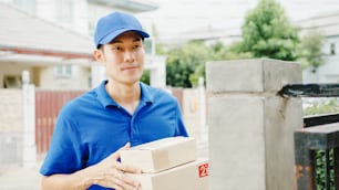 Jeune homme de courrier de livraison postale asiatique en chemise bleue manipulant des boîtes à colis pour l’envoi au client à la maison et femme asiatique reçoivent le colis livré à l’extérieur. Concept de livraison de nourriture pour les achats de colis.