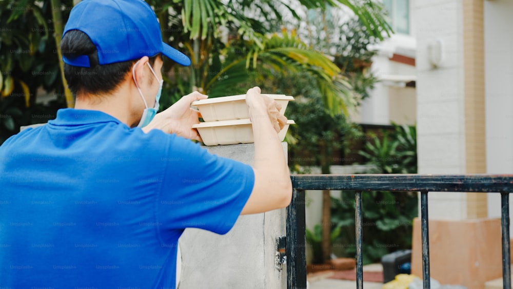 Young postal delivery courier man wear face mask handling food box for send to customer at house and Asian female receive delivered package outdoors. Lifestyle new normal after corona virus concept.