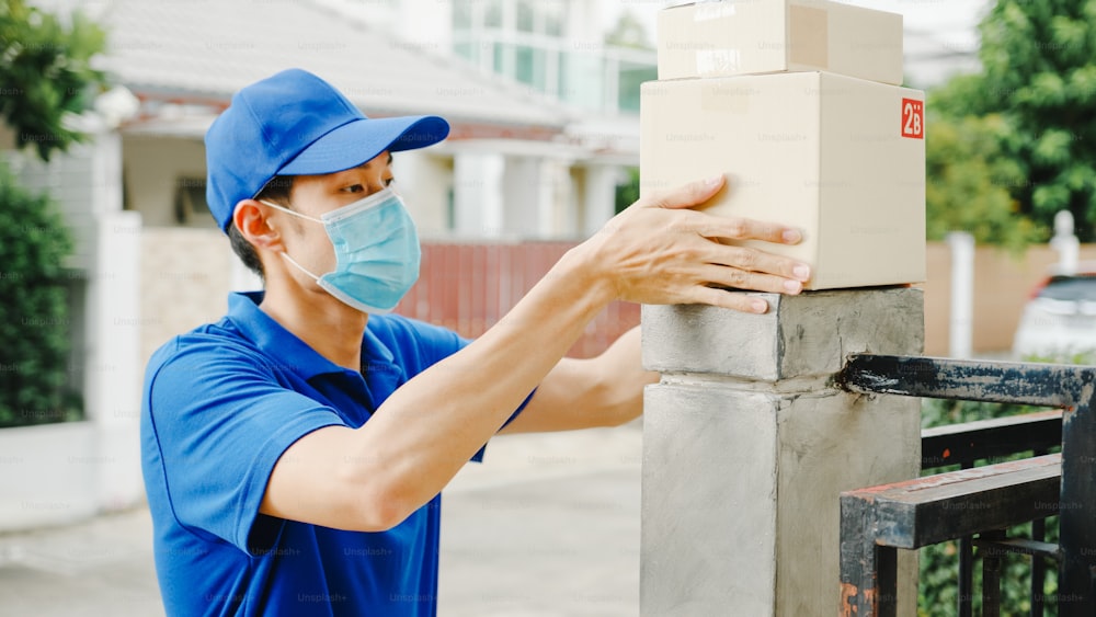 Young postal delivery courier man wear face mask handling parcel box for send to customer at house and Asian female receive delivered package outdoors. Lifestyle new normal after corona virus concept.