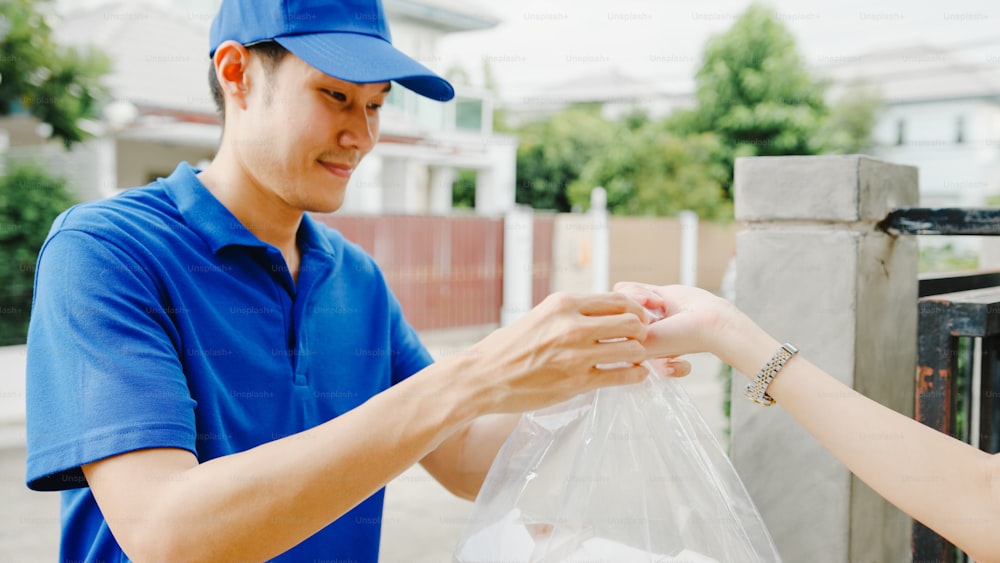 Young Asia postal delivery courier man in blue shirt handling food boxes for sending to customer at house and Asian female receive delivered package outdoors. Package shopping food delivery concept.