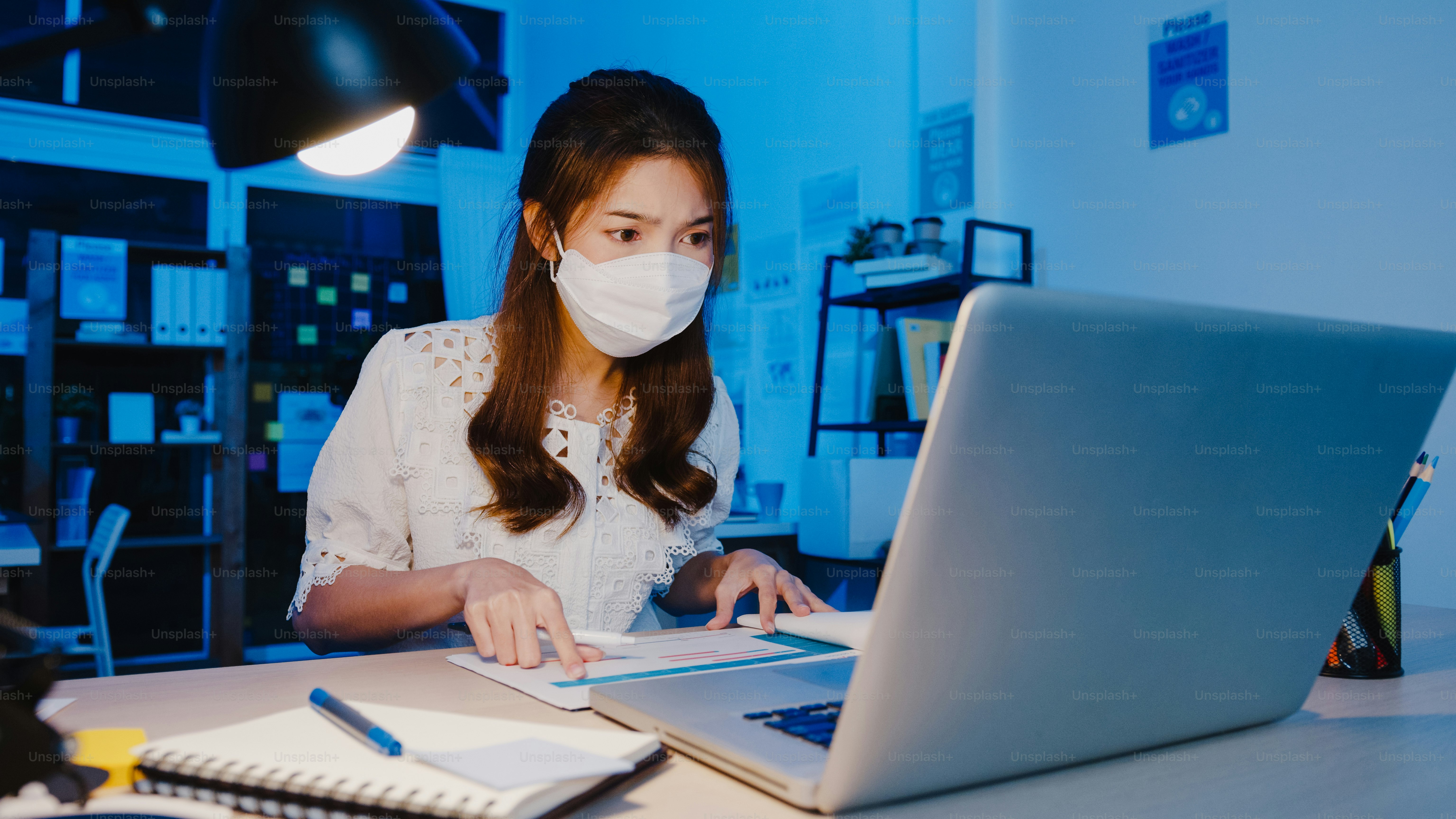 Happy Asia businesswoman wearing medical face mask for social distancing in new normal situation for virus prevention while using laptop back at work in office night. Life and work after coronavirus.