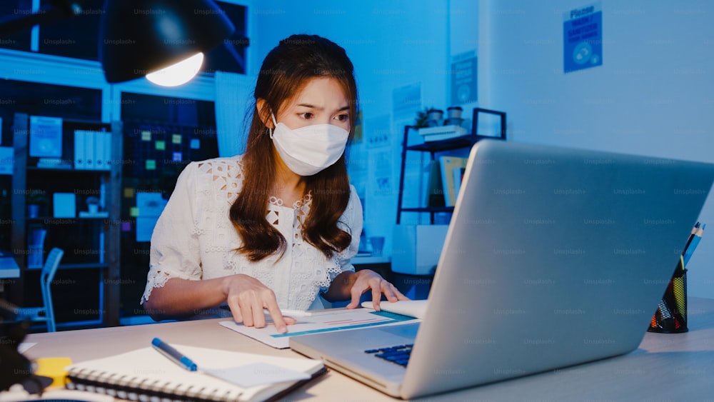 Une femme d’affaires asiatique heureuse portant un masque facial médical pour la distanciation sociale dans une nouvelle situation normale pour la prévention du virus tout en utilisant un ordinateur portable au travail la nuit du bureau. La vie et le travail après le coronavirus.