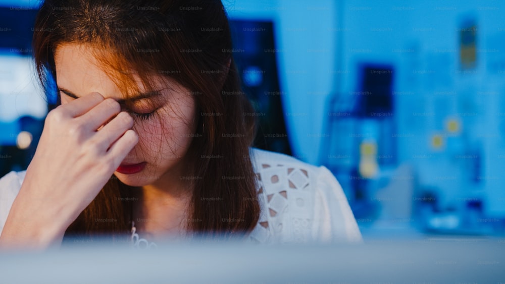 Freelance Asia women using laptop hard work at new normal home office. Working from house overload at night, remotely work, self isolation, social distancing, quarantine for corona virus prevention.