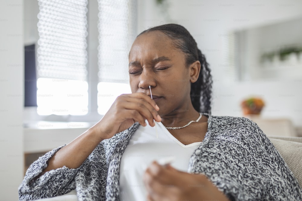 Mujer afroamericana usando un hisopo de algodón mientras se hace la prueba PCR de coronavirus en casa. Mujer usando prueba de diagnóstico rápido de coronavirus. Mujer joven en casa usando un hisopo nasal para COVID-19.