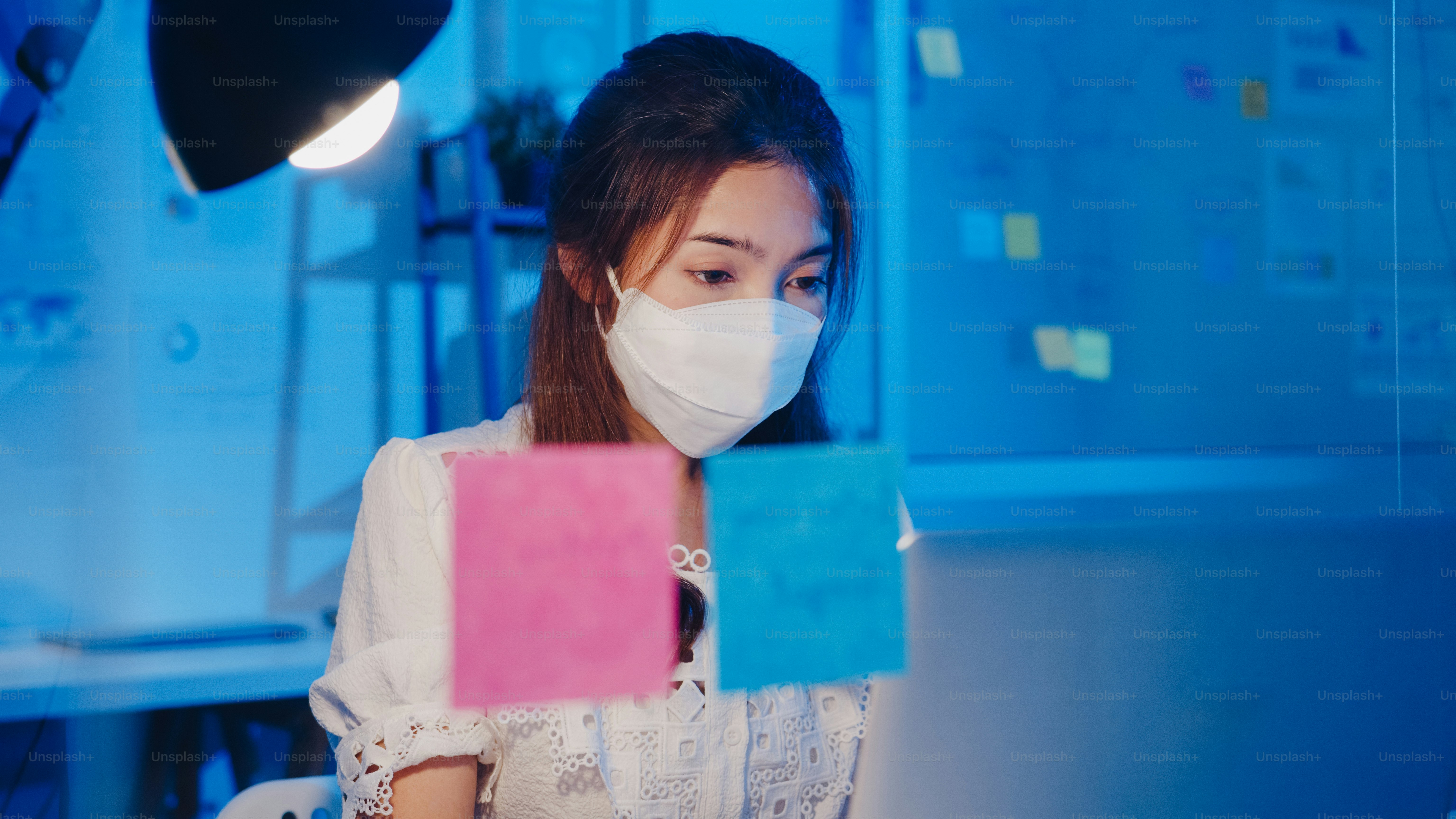 Happy Asia businesswoman wearing medical face mask for social distancing in new normal situation for virus prevention while using laptop back at work in office night. Life and work after coronavirus.