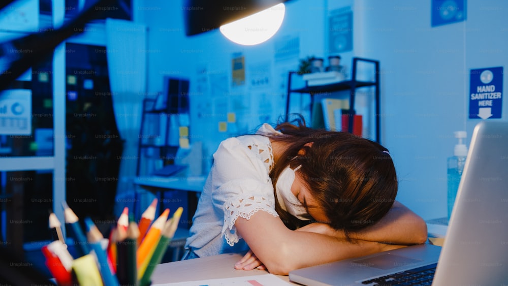 Freelance Asia exhausted lady wear face mask sleeping at new normal home office. Working from house overload at night, Remotely, self isolation, social distance, quarantine for corona virus prevention