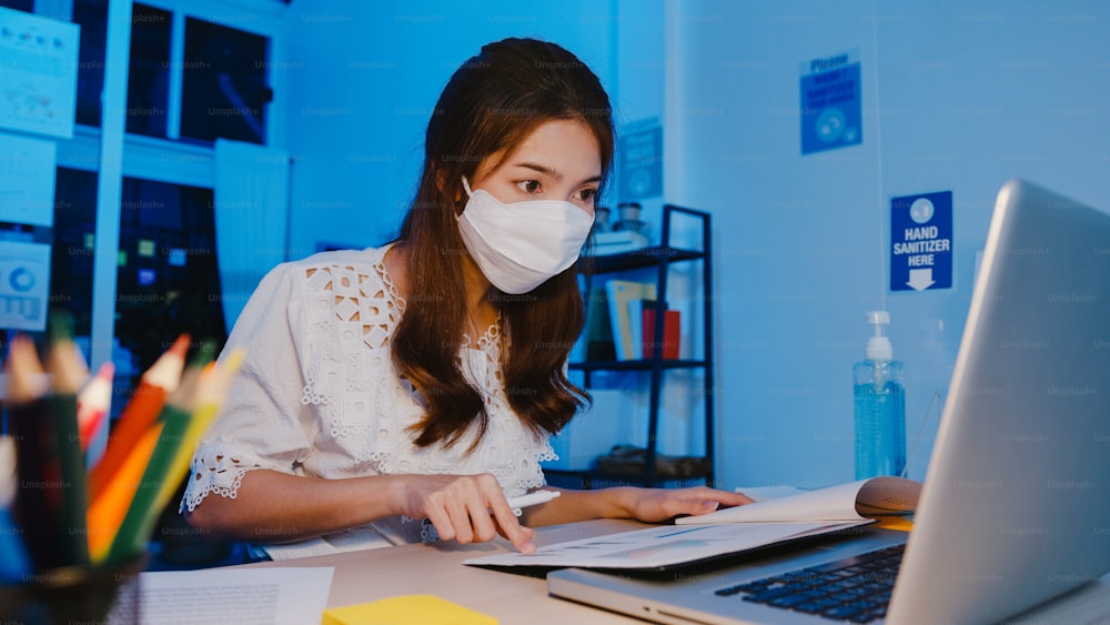 Empresária feliz da Ásia usando máscara facial médica para distanciamento social em nova situação normal para prevenção do vírus enquanto usa laptop de volta ao trabalho no escritório à noite. Vida e obra após o coronavírus.