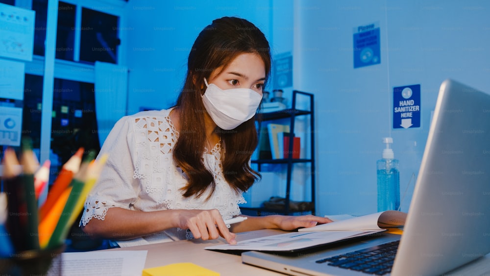 Happy Asia businesswoman wearing medical face mask for social distancing in new normal situation for virus prevention while using laptop back at work in office night. Life and work after coronavirus.