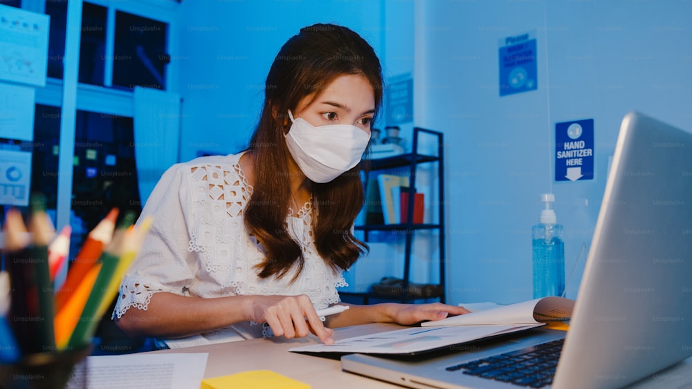 Donna d'affari felice dell'Asia che indossa una maschera facciale medica per il distanziamento sociale in una nuova situazione normale per la prevenzione del virus mentre utilizza il laptop al lavoro nella notte dell'ufficio. Vita e lavoro dopo il coronavirus.