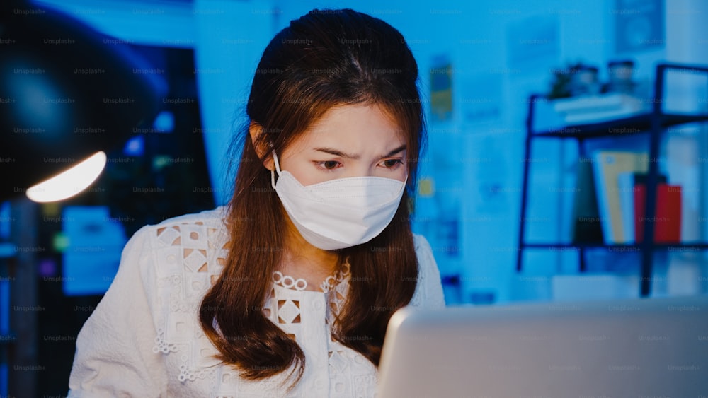 Donna d'affari felice dell'Asia che indossa una maschera facciale medica per il distanziamento sociale in una nuova situazione normale per la prevenzione del virus mentre utilizza il laptop al lavoro nella notte dell'ufficio. Vita e lavoro dopo il coronavirus.