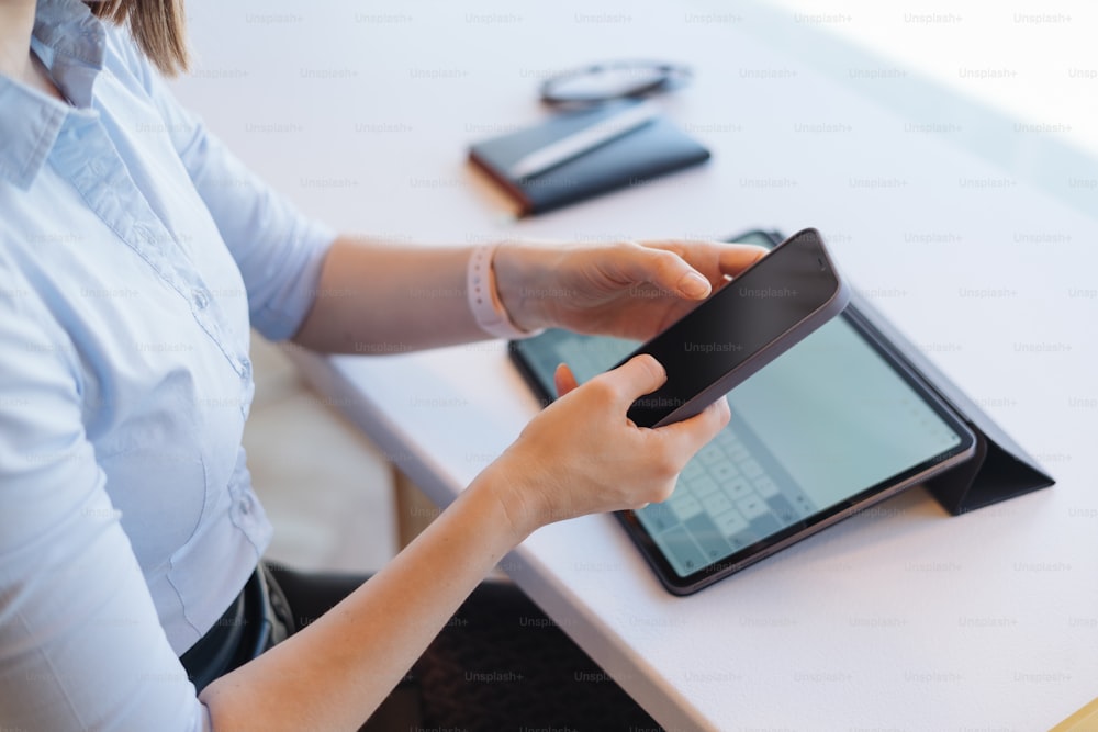 Mani femminili che tengono uno smartphone con schermo nero su sfondo di lavoro, carte, tablet, scrivania bianca