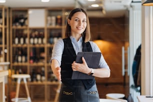 Image of beautiful young woman standing while holding a digital tablet and give her hand. Business owner, cafeteria, restaurant