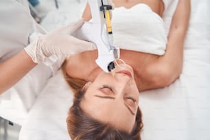 Top view of happy woman relaxing while cosmetologist is using anti age fillers for her face