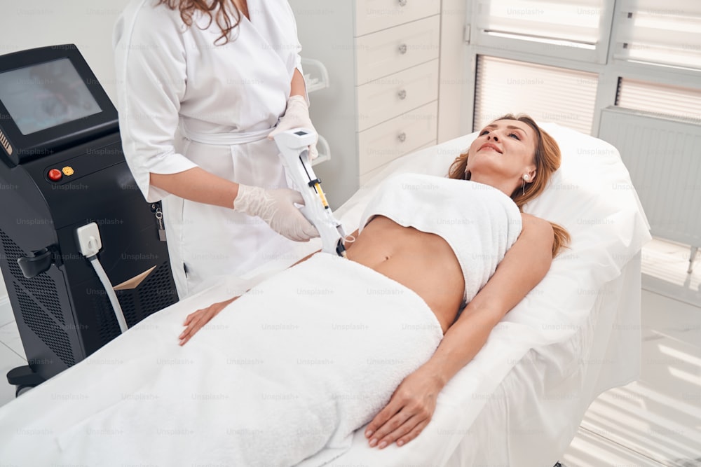 Top view of cheerful pretty female lying on couch while cosmetologist is using laser on her stomach