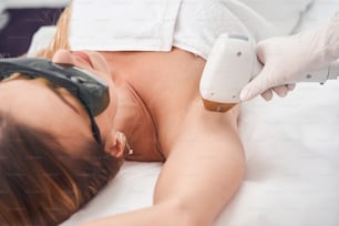 Top view of pretty female lying on couch while professional is using laser device for removing hair
