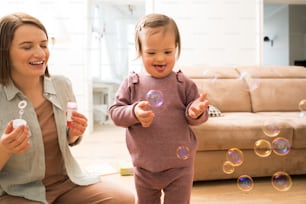 Full length view of the happy girl with down syndrome playing with soap bubbles near her mother at home. Little girl feeling happy. People with special needs concept. Stock photo