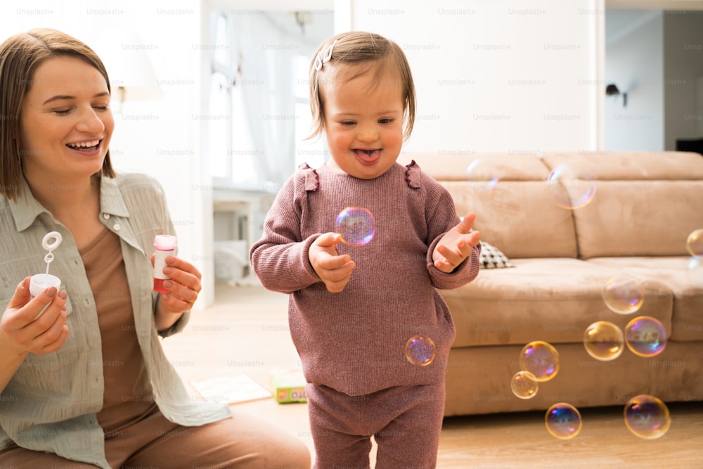 Full length view of the happy girl with down syndrome playing with soap bubbles near her mother at home. Little girl feeling happy. People with special needs concept. Stock photo