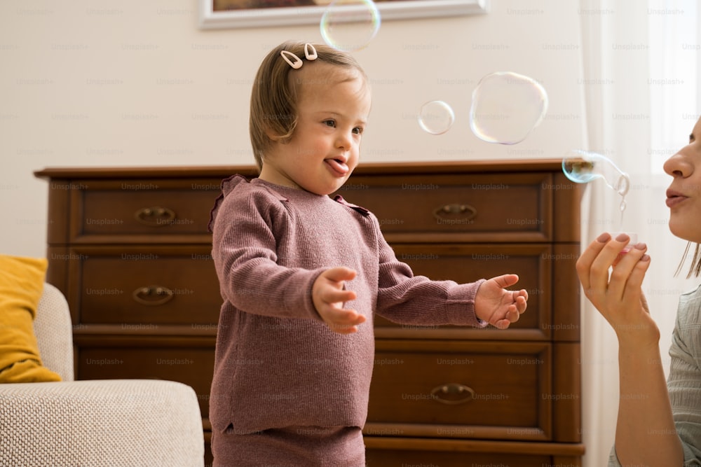 Waist up portrait view of the little girl with down syndrome feeling fun while her young mother blowing soap bubbles at home at the morning. Stock photo