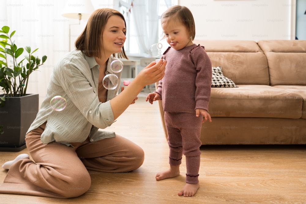 Look at this. Full length view of the cute woman sitting at the floor and showing soap bubbles to her daughter with down syndrome. Stock photo