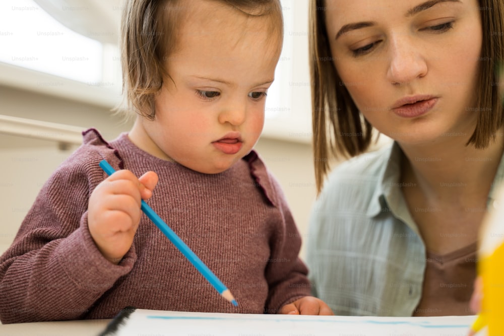 Hmm. Little girl with down syndrome drawing diligently near her mother. Adorable mother telling something to her daughter while she looking at the drawing. People with special needs concept