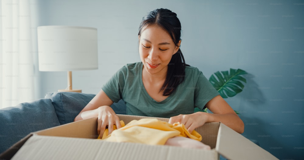 幸せなアジアの女性は段ボール箱のパッケージをエキサイティングに開き、自宅のリビングルームでオンライン市場から高品質のファッション布製品を試してマッチングして楽しんでください。オンラインショッピングと配送のコンセプト。