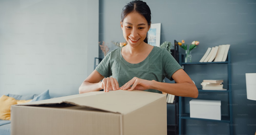 Happy beautiful Asia lady unboxing cardboard delivery package from online marketplace in living room at house. Satisfied shopper in internet unboxing goods, Online shopping and delivery concept.