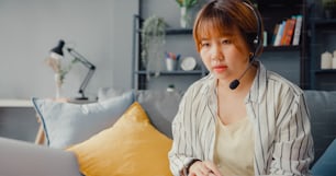 Empresaria asiática que usa una computadora portátil habla con colegas sobre el plan en una videollamada mientras trabaja desde casa en la sala de estar. A distancia en el lugar de trabajo, distanciamiento social, cuarentena para la prevención del coronavirus.
