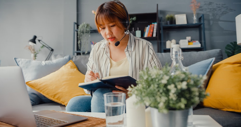 Asia businesswoman using laptop talk to colleagues about plan in video call while working from house at living room. Remotely at workplace, social distancing, quarantine for corona virus prevention.