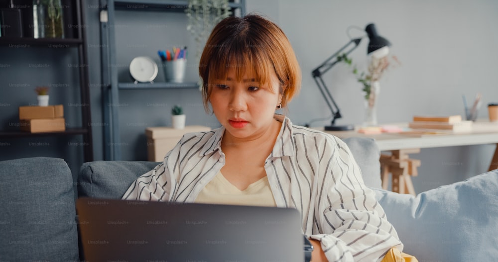 Freelance Asia lady feel headache while sitting on couch with laptop online learn in living room at house. Working from home, remotely work, social distancing, quarantine for corona virus prevention.