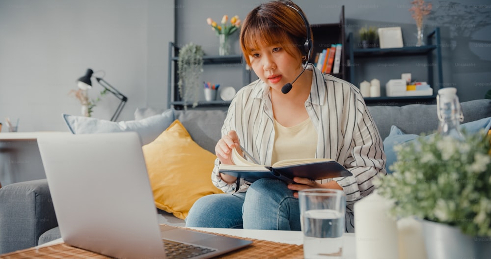 Empresaria asiática que usa una computadora portátil habla con colegas sobre el plan en una videollamada mientras trabaja desde casa en la sala de estar. A distancia en el lugar de trabajo, distanciamiento social, cuarentena para la prevención del coronavirus.