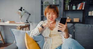 Giovane signora asiatica che usa la videochiamata dello smartphone parla con la famiglia sul divano nel soggiorno di casa. Lavorare da casa da remoto durante il blocco covid-19, il distanziamento sociale, la quarantena per la prevenzione del coronavirus.