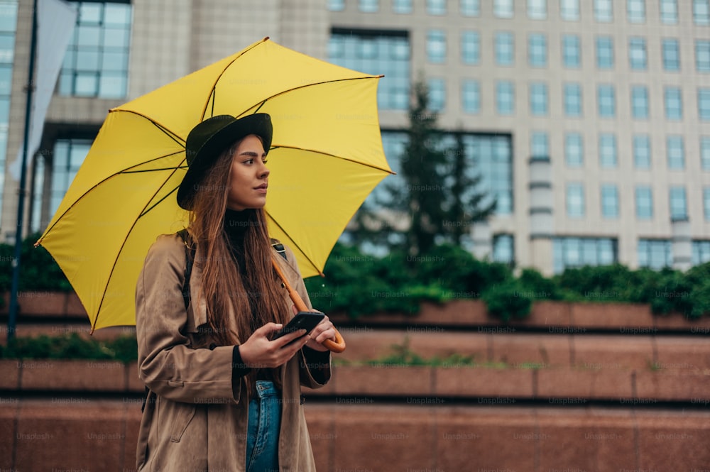 スマートフォンを使い、雨の日に外で黄色い傘を持つ若い美しい女性
